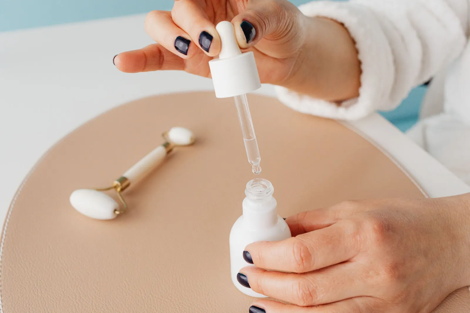 Dłonie nabierają serum pipetą z buteleczki, obok leży roller do masażu twarzy.